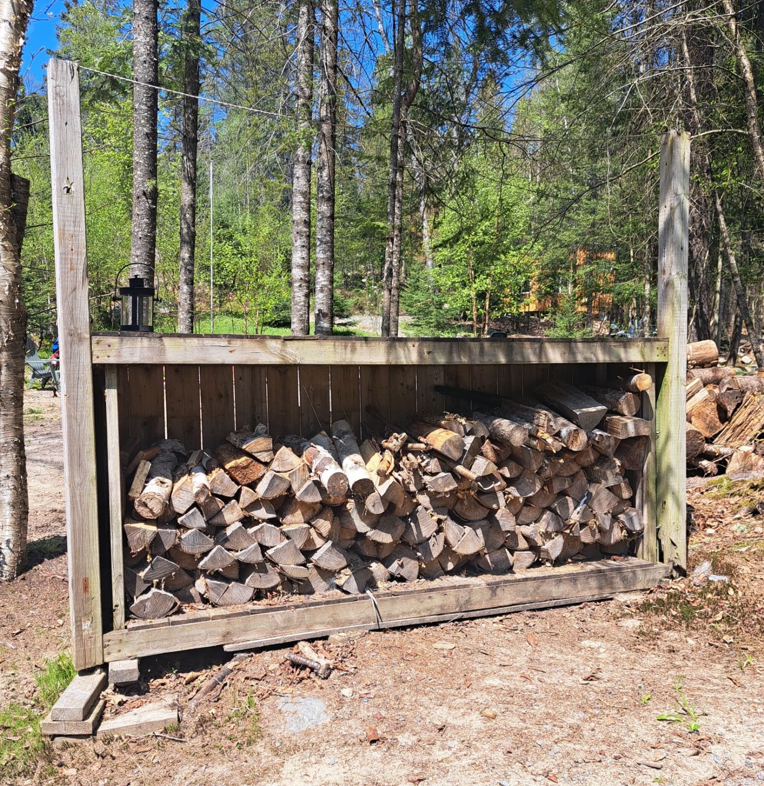 A wooden storage rack filled with stacked firewood, set in a forested area with trees and greenery.