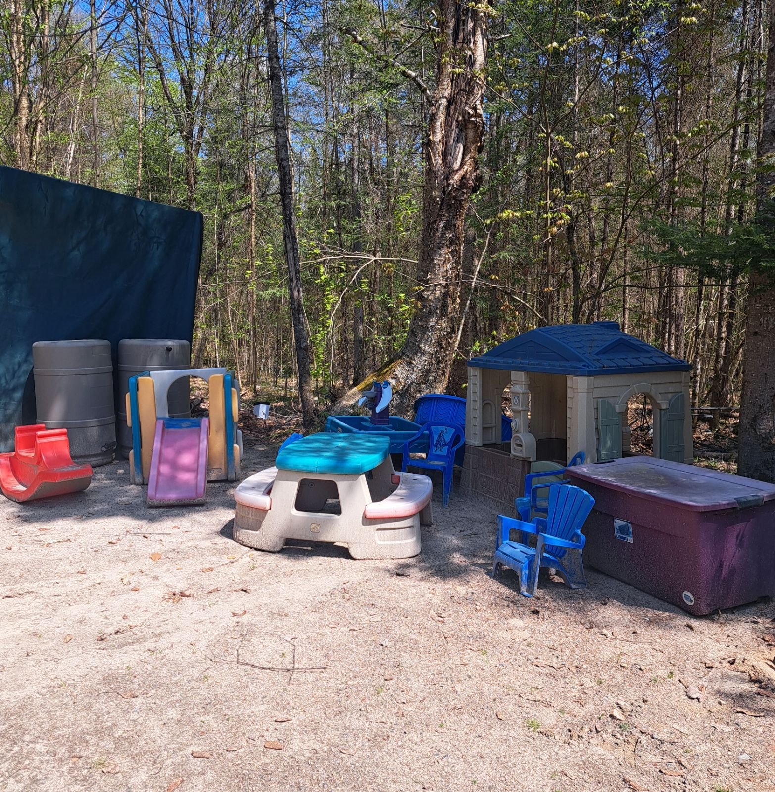 A children's play area with various plastic play structures, including a small house, table, and chairs. The play area is set on sandy ground and surrounded by trees.