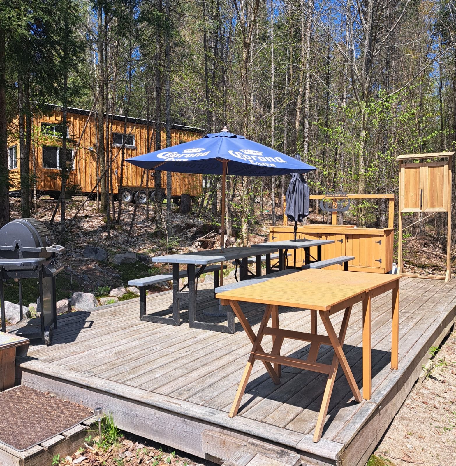 A wooden deck area with picnic tables, a BBQ grill, and a Corona umbrella providing shade. The deck is surrounded by trees and there is a small building in the background.