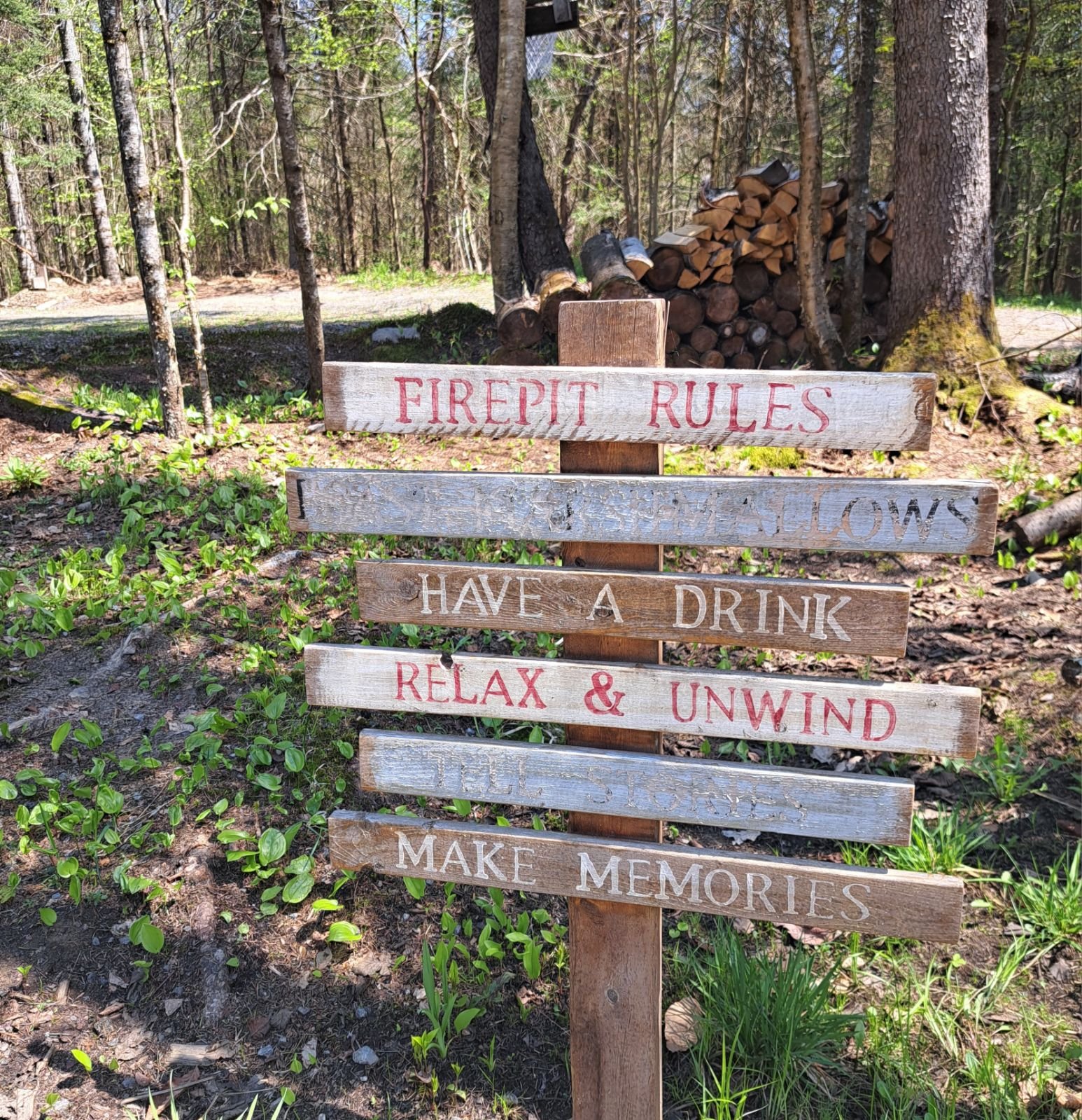 A wooden sign with firepit rules written on it. The rules include "Firepit Rules," "No Shoes on Firewood," "Have a Drink," "Relax & Unwind," and "Make Memories." The sign is located in a wooded area.